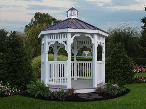 Small octagonal white vinyl gazebo with a bronze roof sitting in a flowerbed