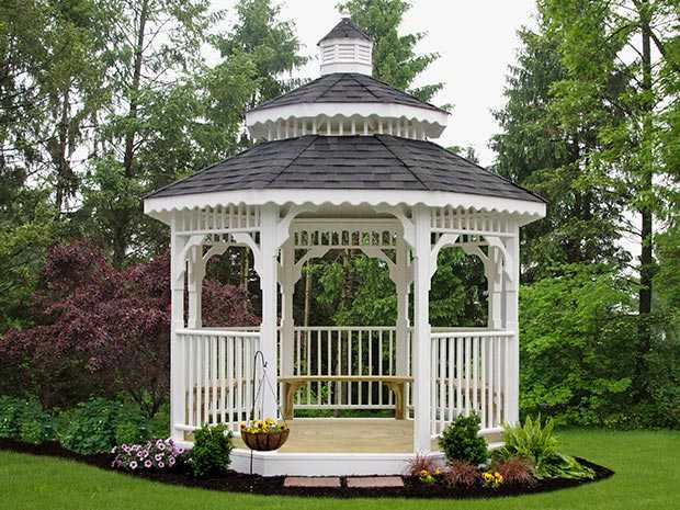 A beautiful White octagon gazebo with benches, a double shingled roof, and a pretty cupola on top nestled in some trees.